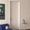 An elegant off-white paneled interior door with an arched top section and a gold handle, set within a classic cream-colored room featuring molding, a blue velvet armchair, and a small side table.