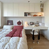 A modern bedroom interior featuring a built-in white desk and shelving unit under cream-colored wall cabinets. A bed with patterned bedding and a red checkered throw is on the left, and a white chair with black legs sits at the desk.