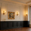 A large interior room scene with cream paneled walls, black wainscoting, warm wall sconces, a framed landscape painting, dark wood flooring, and a window with blinds. Smaller inset thumbnails along the bottom show mirrored or repeated views of the same interior design.