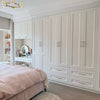 A large white built-in wardrobe and vanity cabinet system in a styled bedroom, featuring paneled doors, drawers, and a built-in mirror. A pink bed and light wood flooring are visible in the foreground.