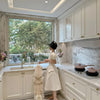 A woman in a cream-colored dress stands in a bright, modern kitchen with white cabinets and marble backsplash, holding a fluffy white dog near the sink window.