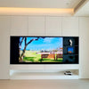 A modern wall-mounted television display set in a minimalist white interior, featuring a large screen showing a park scene and a smaller side display shelf.