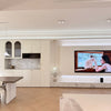A modern living room interior with a large wall-mounted television displaying a scene, set above a marble-topped console. A kitchen area with cream cabinets and a marble backsplash is visible on the left, alongside a dining table and a beige sofa in the foreground.